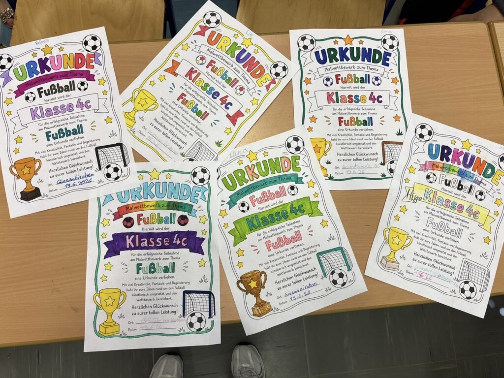 Auf einem Schreibtisch liegen sechs bunte Urkunden für ein Fußballturnier der Klasse 4c ausgebreitet. Jede Urkunde ist mit Zeichnungen von Pokalen, Fußbällen und Ziertext verziert und enthält handgeschriebene Namen und Angaben.