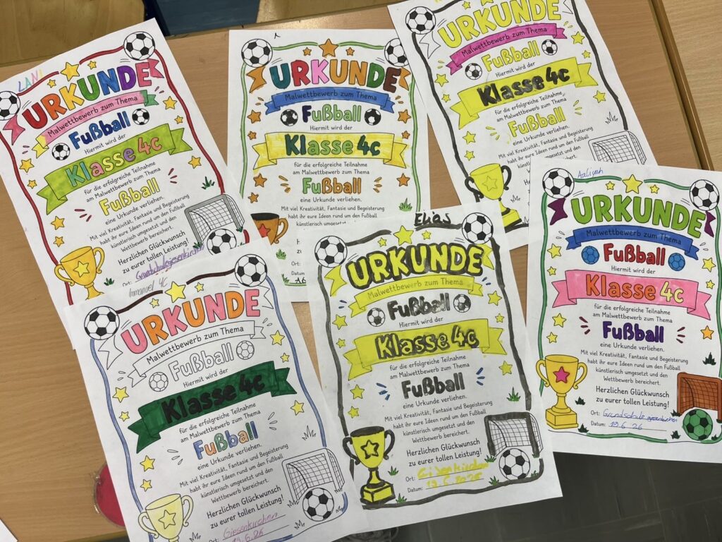 Auf einem Holztisch liegen sechs bunte, handbemalte Urkunden für eine Fußballveranstaltung in der Klasse 4c, die jeweils mit Zeichnungen von Fußbällen, Pokalen und Toren verziert sind.