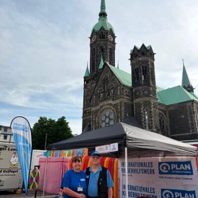 Zwei Personen stehen lächelnd vor einem Stand von Plan International bei einer Veranstaltung im Freien. Hinter ihnen steht eine historische Kirche mit einem hohen Kirchturm und grünen Dächern. Der Stand ist mit Bannern, einer Regenbogenflagge und Infotafeln ausgestattet.