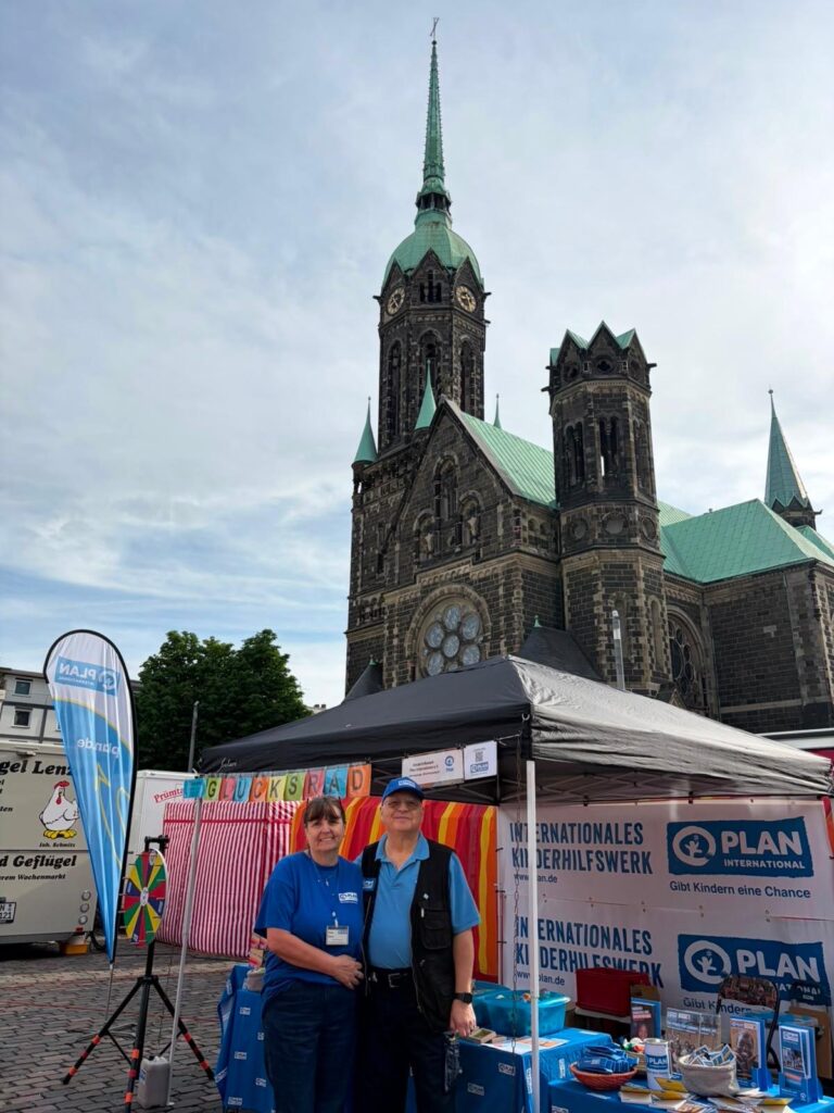 Zwei Personen stehen lächelnd vor einem Stand von Plan International bei einer Veranstaltung im Freien. Hinter ihnen steht eine historische Kirche mit einem hohen Kirchturm und grünen Dächern. Der Stand ist mit Bannern, einer Regenbogenflagge und Infotafeln ausgestattet.