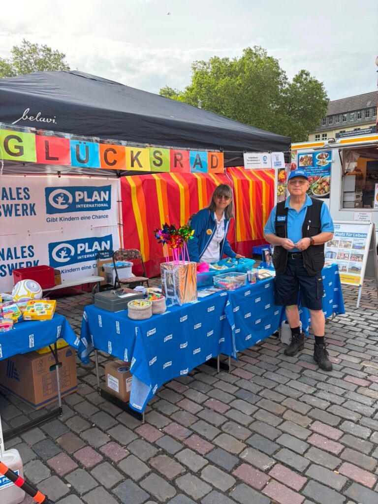 Zwei Personen stehen hinter einem Stand mit einem blauen Tischtuch und bunten Preisen. Auf einem Schild darüber steht "GLÜCKSRAD". Der Stand wirbt für Plan International und ist auf Kopfsteinpflaster im Freien unter einem Zelt aufgebaut.