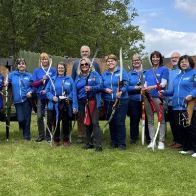 Eine Gruppe von dreizehn Personen in blauen Hemden steht auf einer Wiese vor Bogenschießscheiben, Pfeil und Bogen in der Hand, und lächelt an einem sonnigen Tag in die Kamera, während hinter ihnen Bäume und ein Netz zu sehen sind.