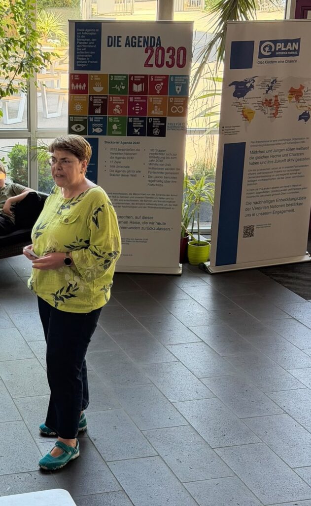 Eine Frau in einem gelben Shirt spricht in einem Innenraum vor Bannern, die sich auf die Agenda 2030 und eine PLAN-Initiative beziehen. Sie steht auf grauen Fliesen, im Hintergrund sind Sonnenlicht und Pflanzen zu sehen.