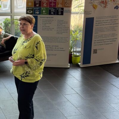 Eine Frau in einem gelben Oberteil spricht in einem Raum vor Plakaten zur Agenda 2030 und zu Plan International. Sonnenlicht fällt durch die Fenster, und im Hintergrund sind Pflanzen zu sehen.
