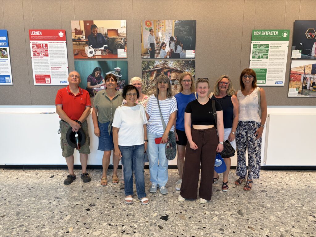 Zehn Personen stehen in einem Raum vor einer Wand, an der Informationsplakate und Fotos angebracht sind. Die Gruppe ist leger gekleidet, lächelt und posiert gemeinsam auf einem gesprenkelten Fliesenboden.