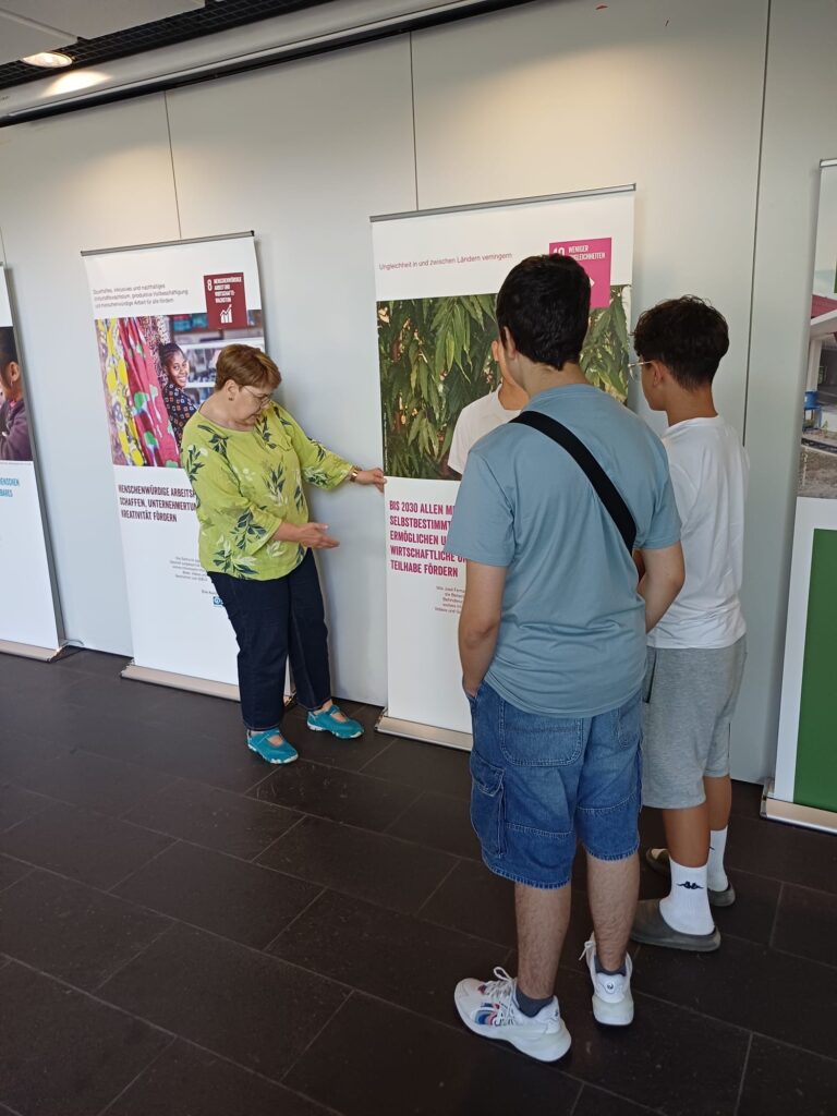 Eine Frau deutet auf eine Informationstafel, während sie mit drei Jungen spricht, die vor ihr in einem hell beleuchteten Innenraum stehen.