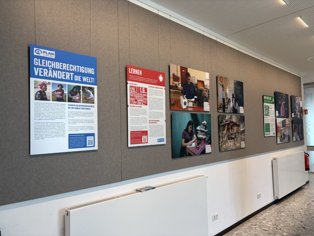 An einer Wand im Flur sind Plakate und Fotos zum Thema Gleichberechtigung und Bildung ausgestellt, mit deutschen Texten, Personenbildern und Informationsgrafiken an einer grauen Wand über weißen Heizkörpern.