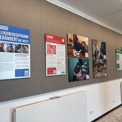 An einer Wand im Flur sind Plakate und Fotos zum Thema Gleichberechtigung und Bildung ausgestellt, mit deutschen Texten, Personenbildern und Informationsgrafiken an einer grauen Wand über weißen Heizkörpern.