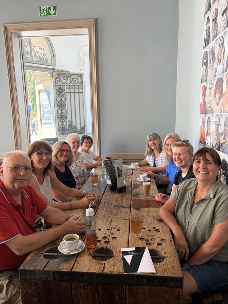 Eine Gruppe von neun Personen sitzt in einem Café um einen Holztisch herum und lächelt in die Kamera. Auf dem Tisch stehen Getränke, und an der Wand hängen gerahmte Fotos. Sonnenlicht fällt durch ein Fenster herein, und die Wände des Cafés sind hell gestrichen.