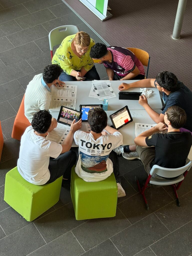 Sechs Personen sitzen um einen Tisch herum und arbeiten gemeinsam mit Unterlagen und Tablets. Bunte Stühle umgeben die Gruppe, und im Mittelpunkt scheint ein gemeinsames Projekt oder eine Lernsitzung zu stehen.