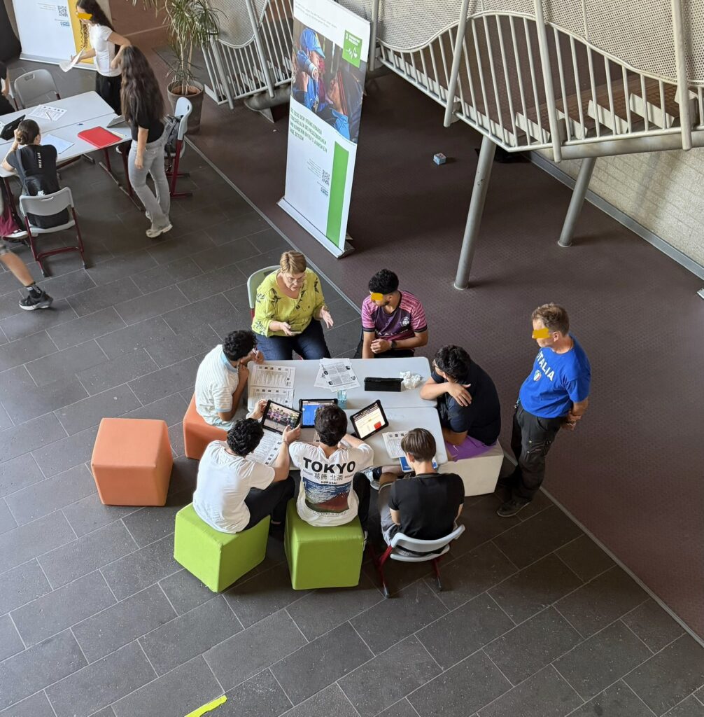 Eine Gruppe von Menschen sitzt mit Laptops und Unterlagen um einen Tisch herum und arbeitet gemeinsam an einem Projekt in einem modernen Innenraum, während andere in der Nähe stehen und zuschauen.